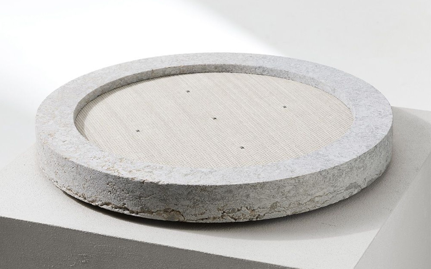 Pigna by POQQ, a round natural stone wireless charger with a woven leather top, resting on a pale plinth in soft daylight
