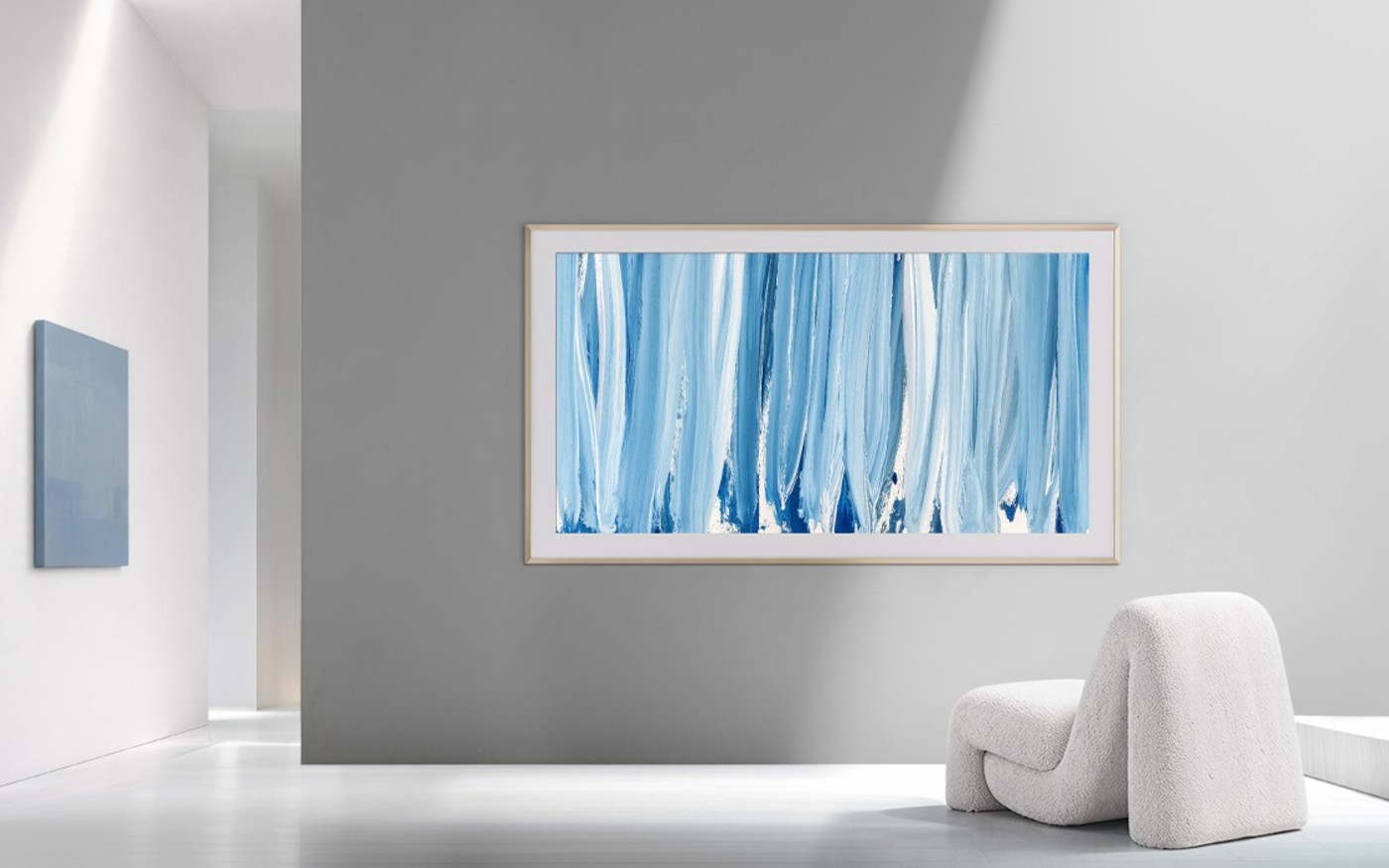 Samsung The Frame Pro hung on a pale grey wall, showing an abstract blue painting, beside a white lounge chair